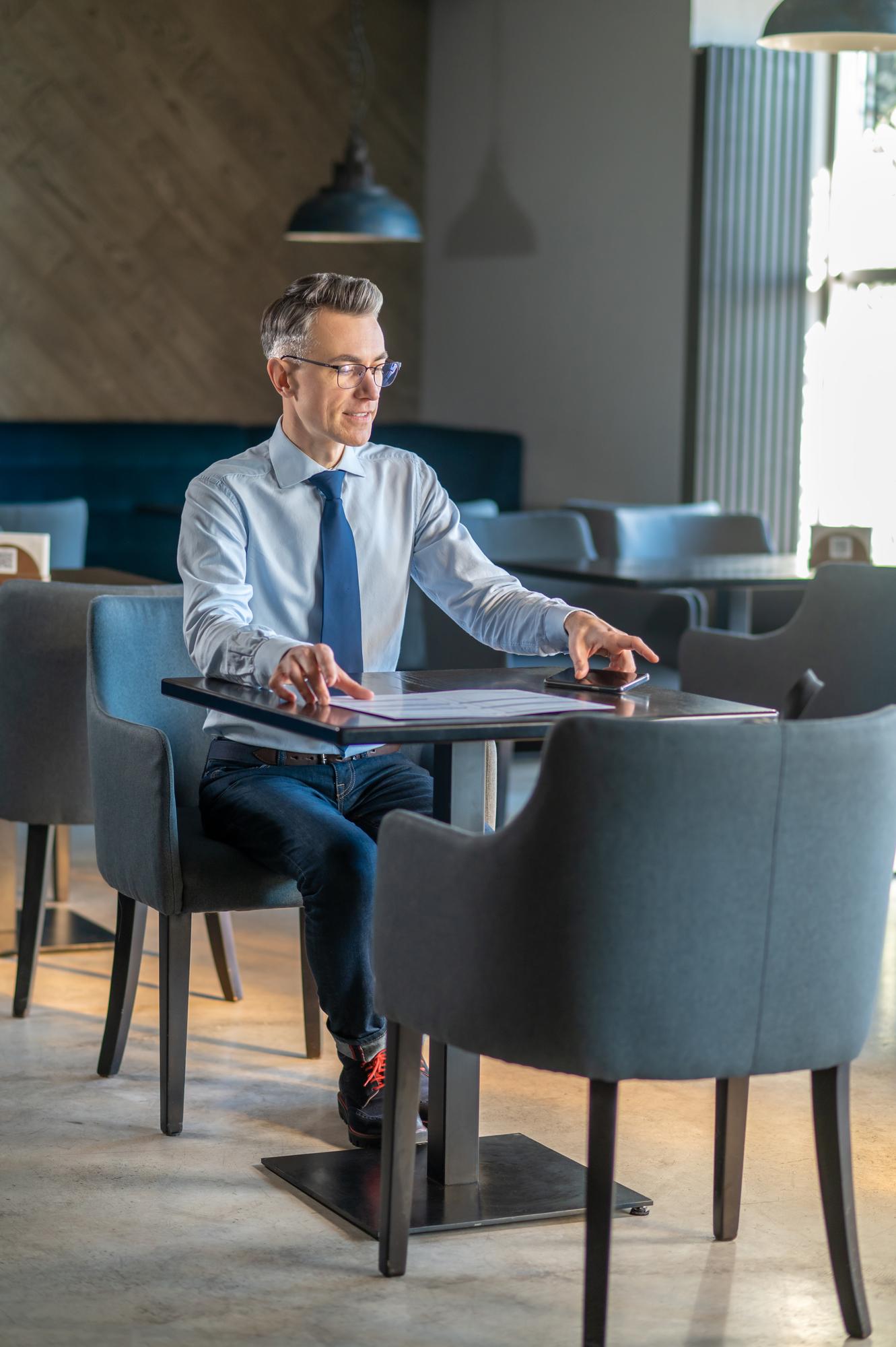Ein Mann mit Krawatte sitzt allein an einem kleinen Tisch in einem caféähnlichen Raum, vor ihm ein Dokument und ein Telefon.